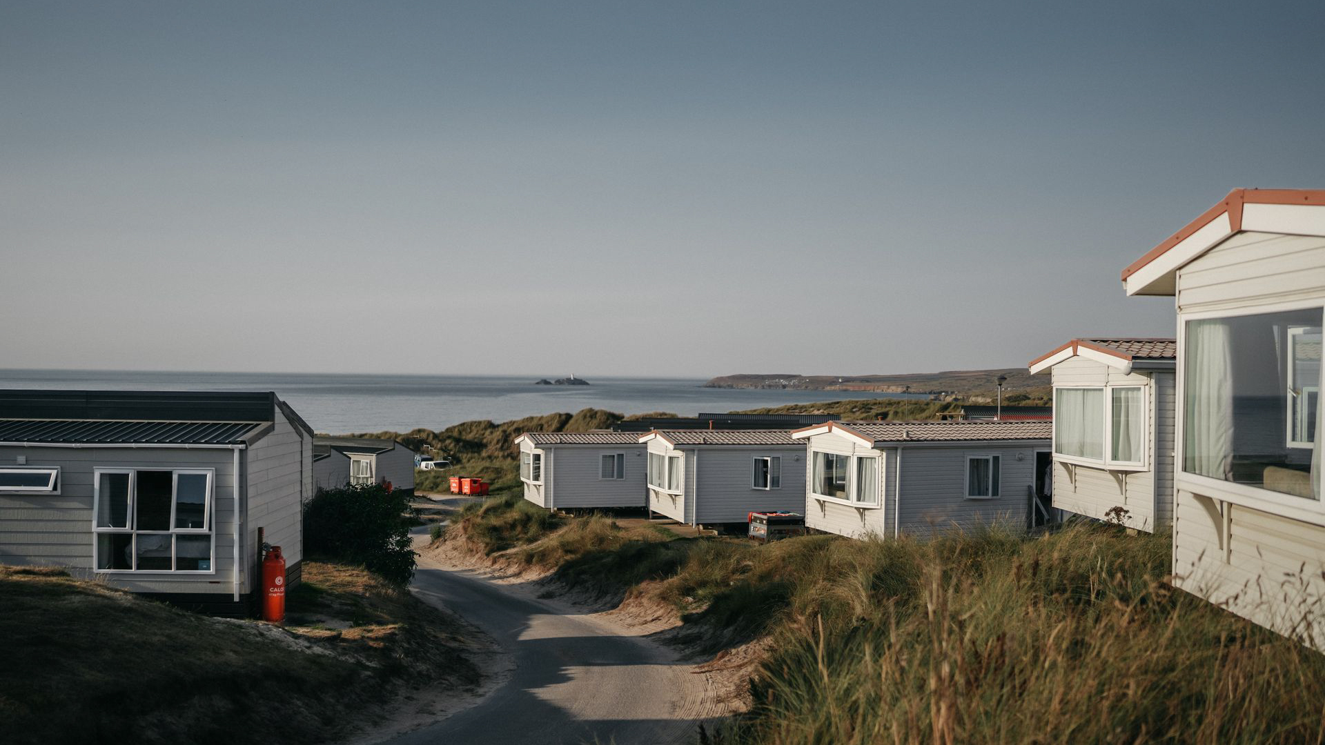 Row of white static caravans on grassy sand dunes overlooking a calm sea at sunset.