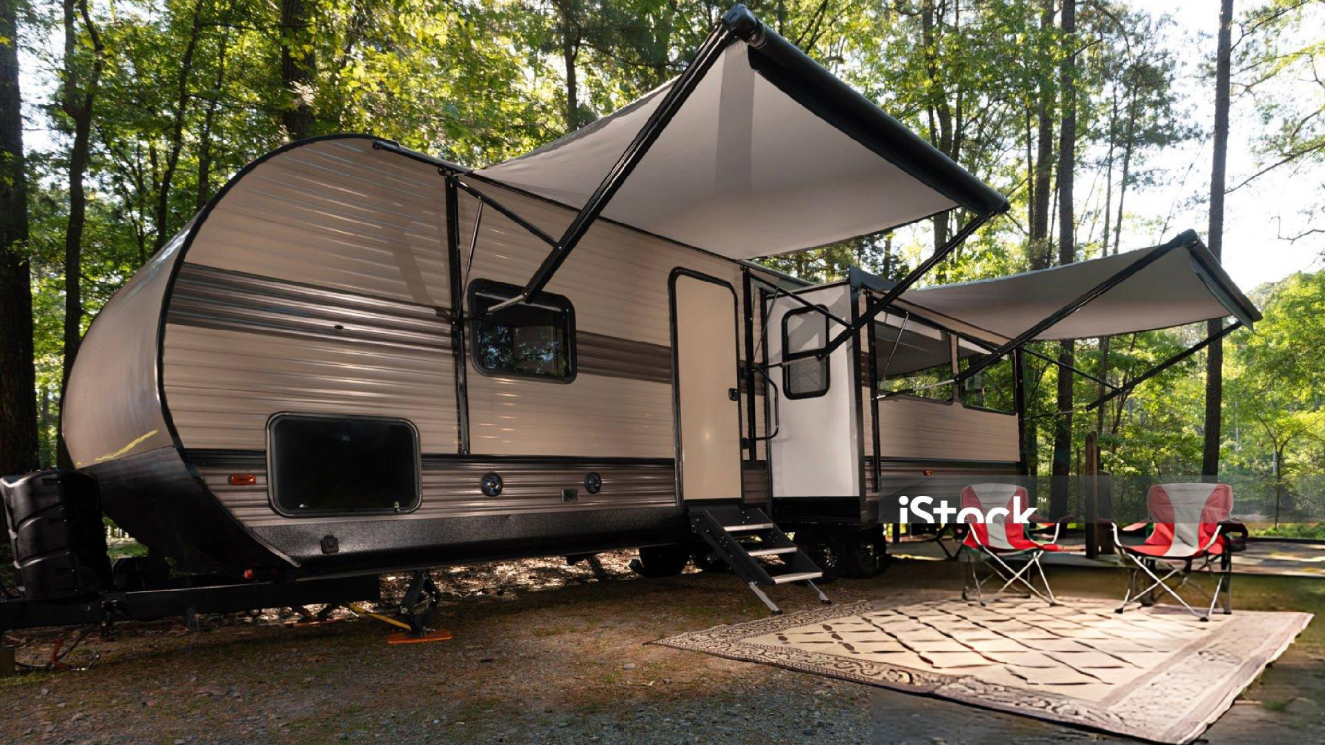 Travel trailer with open awnings parked in a wooded campsite, with two red camping chairs outside.