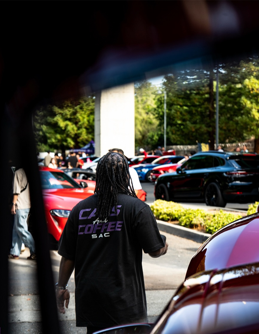 Attendee in a Cars and Coffee Sac shirt seen from behind looking out over parked cars