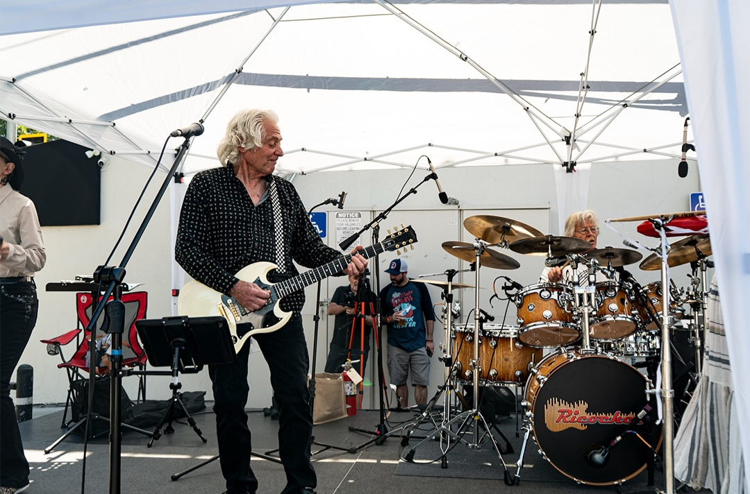 Band performing live under a canopy at a Cars and Coffee Sac event guitarist and drummer visible