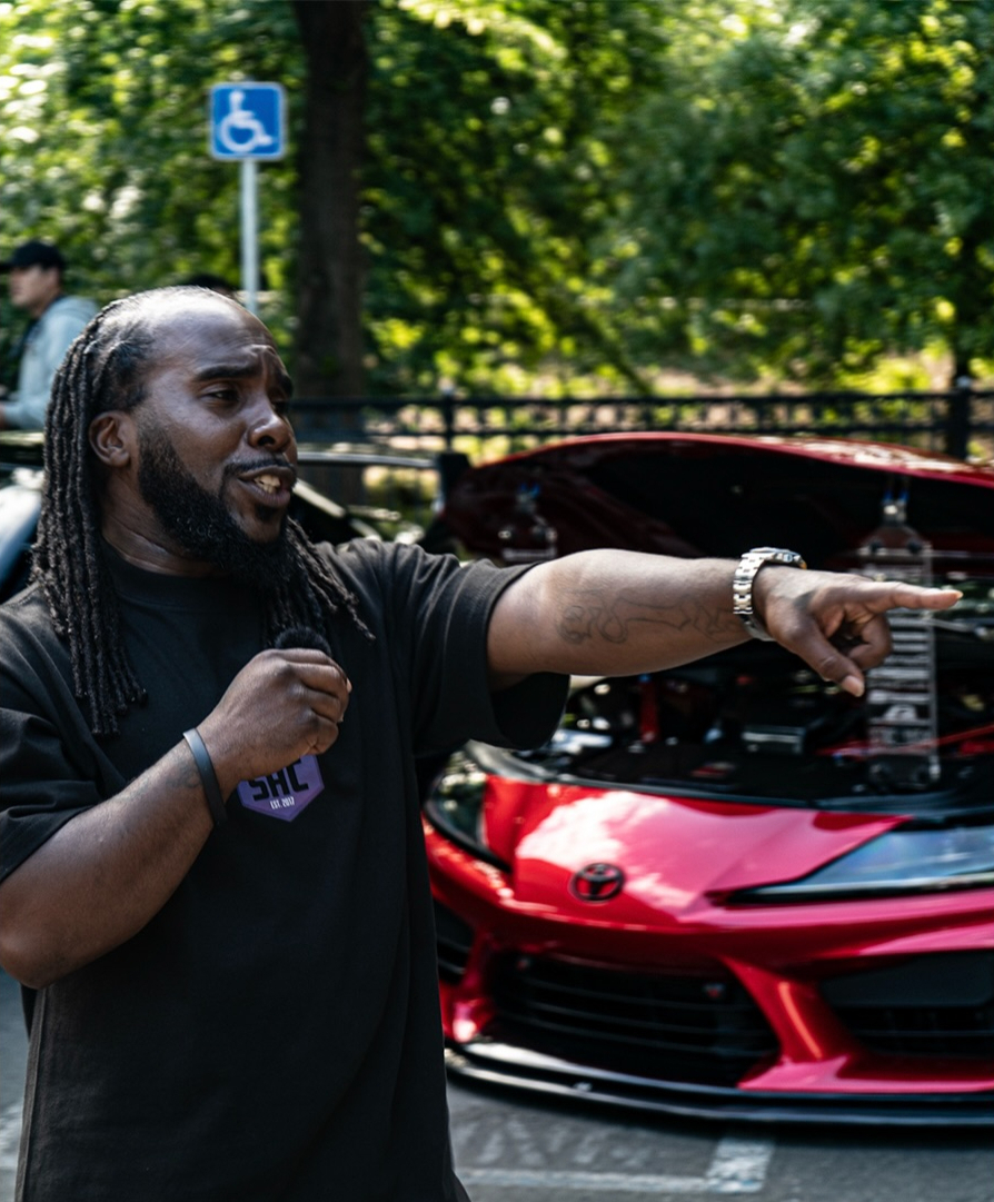Attendee gesturing beside a red car with its hood raised at an event