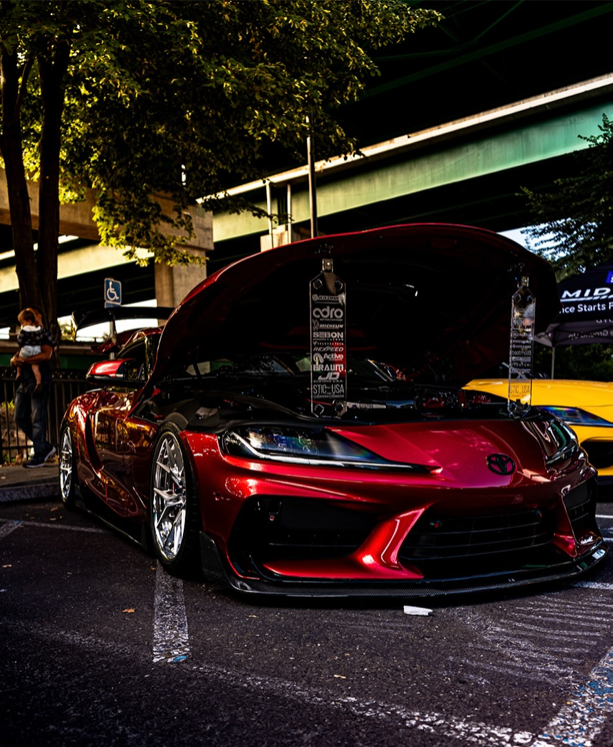 Red car with its hood raised at an event under trees at dusk
