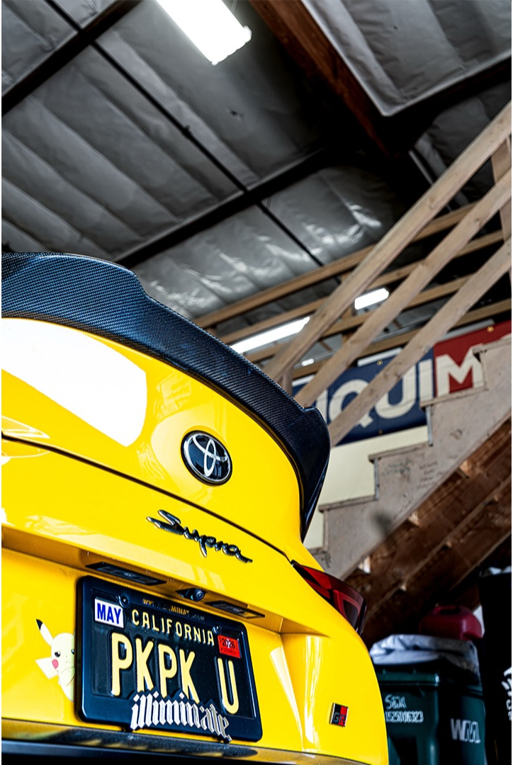 Rear deck and spoiler of a yellow Toyota Supra under workshop lighting
