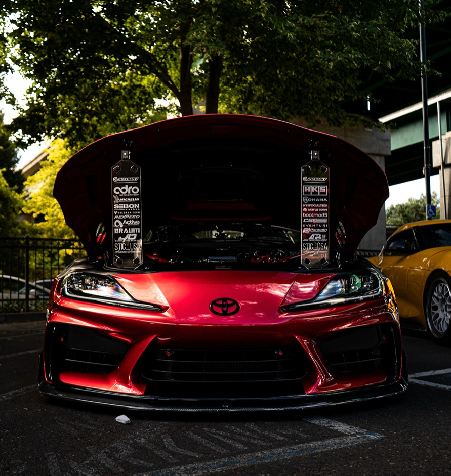 Red sports coupe with its hood raised and display placards propped underneath parked under trees