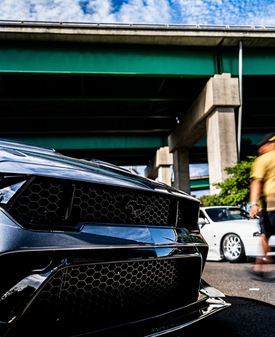 Front of a black Mustang parked under a freeway overpass as attendees walk past