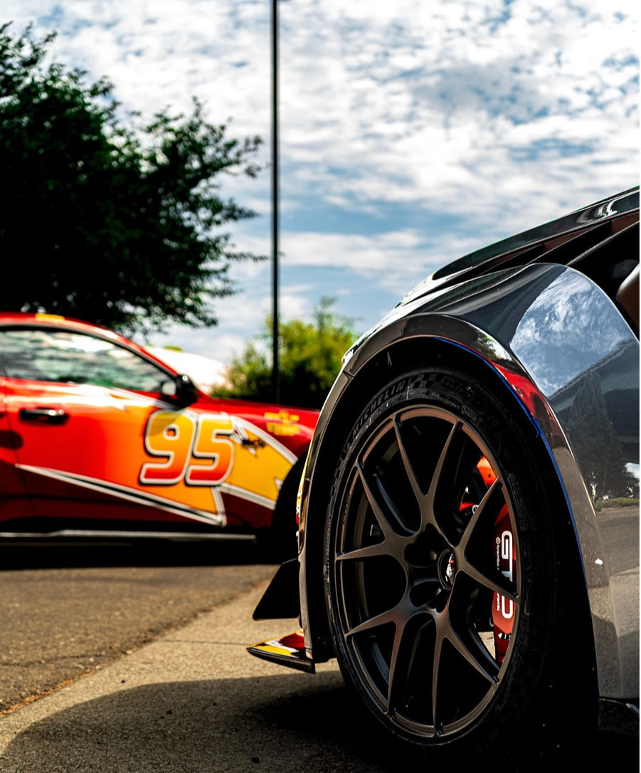 Rear wheel and wide fender of a dark car at an event with a red number 95 race liveried car behind it