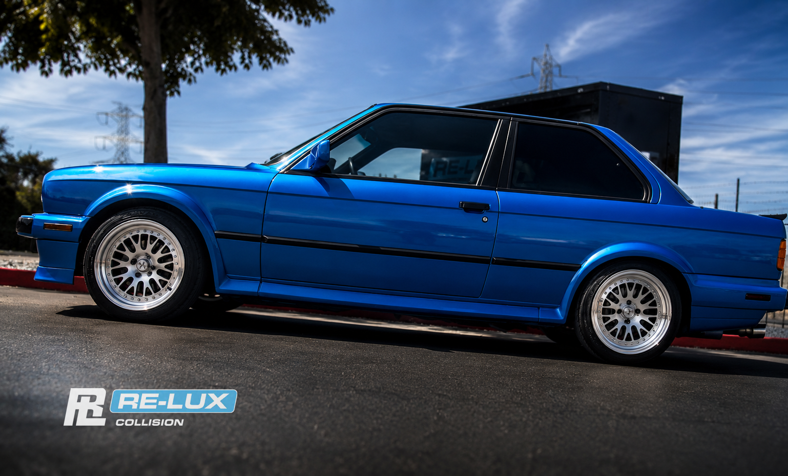 Blue BMW 3 Series, side profile, parked in front of the Re-Lux Collision sign