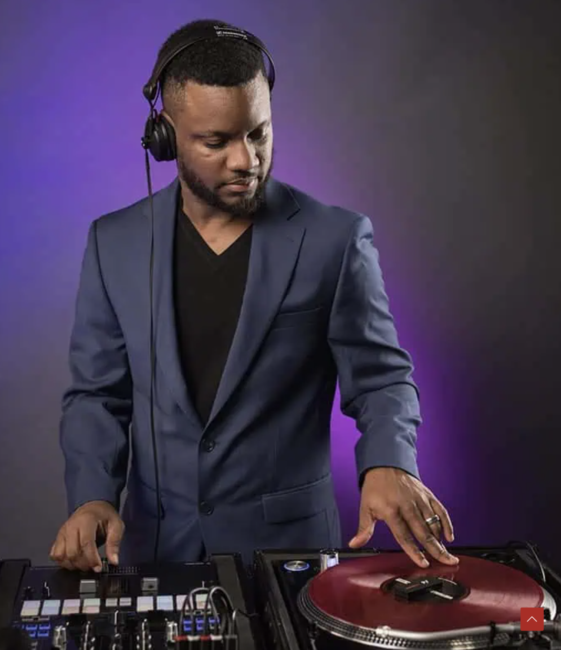 A man in a blue blazer and headphones operates DJ equipment, adjusting a red turntable against a purple-lit background.