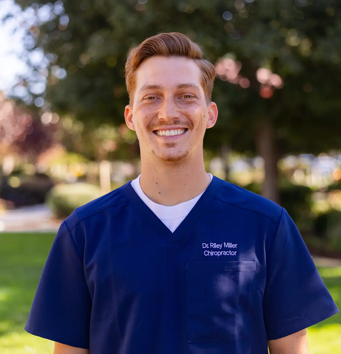 Dr. Riley Miller, chiropractor at Miller Chiropractic and Wellness in Alameda, smiling outdoors in a blue uniform, specializing in spine health and patient care.