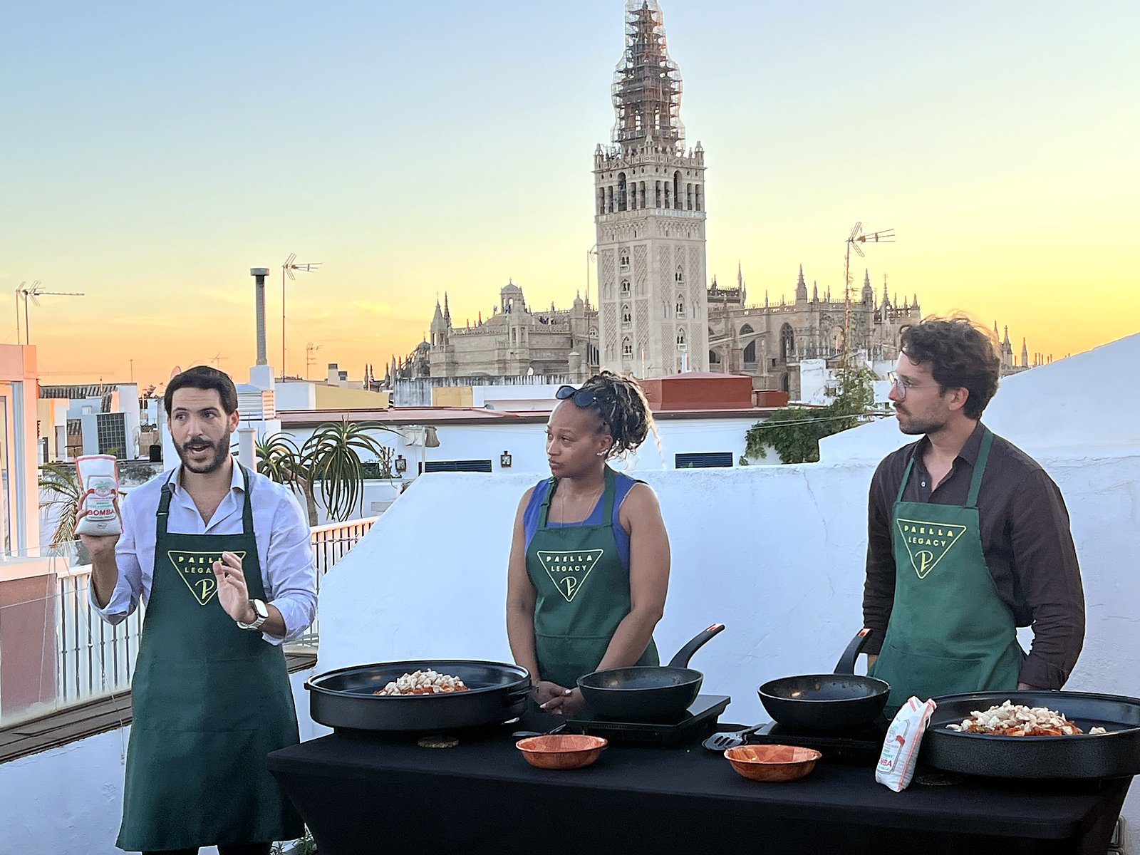 Fernando, in a Paella Legacy apron, guiding two guests at their paella pans on the rooftop, the Giralda behind them