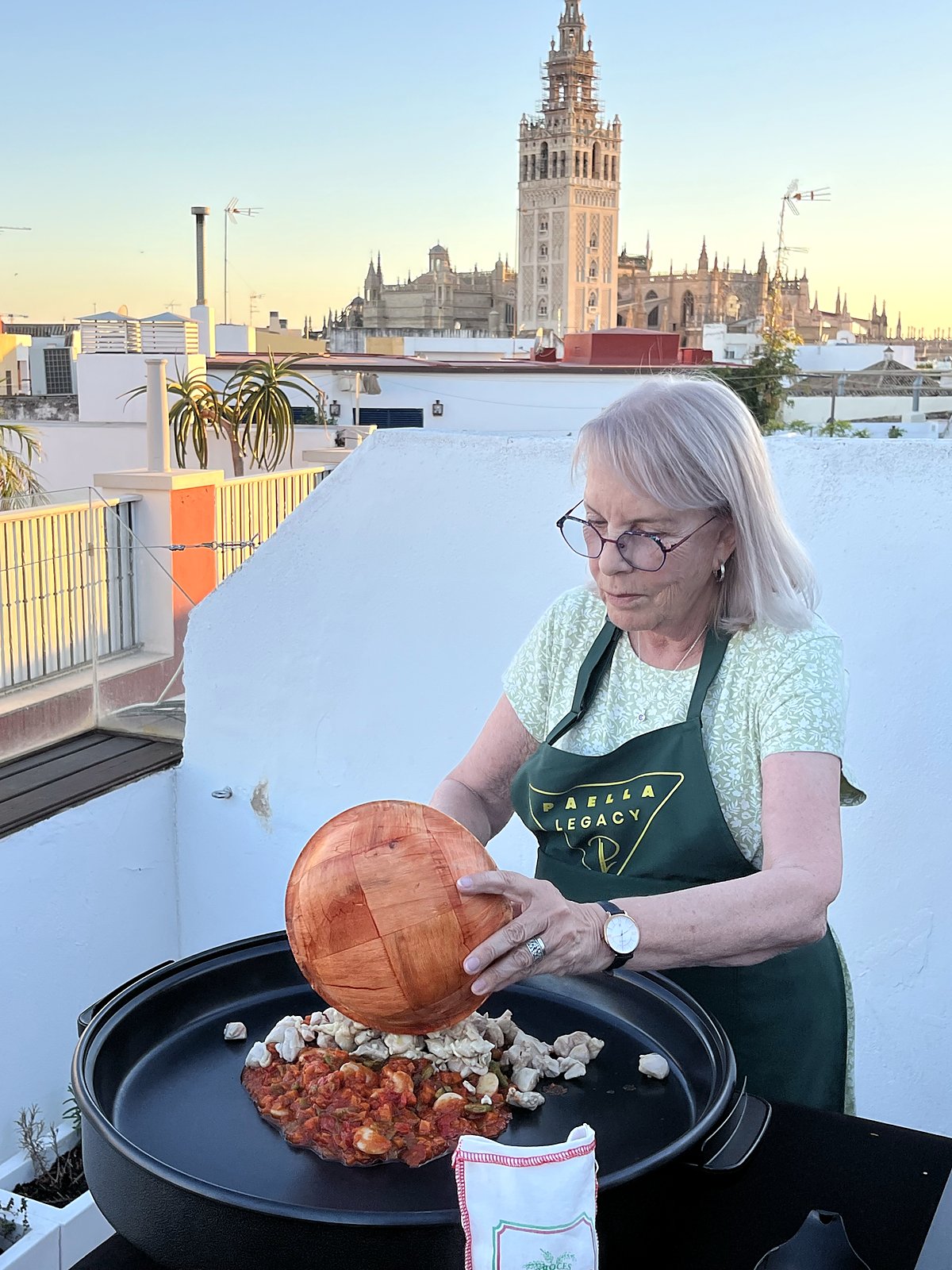 A guest at a Paella Legacy class tipping ingredients into the paella pan, the Giralda in the background
