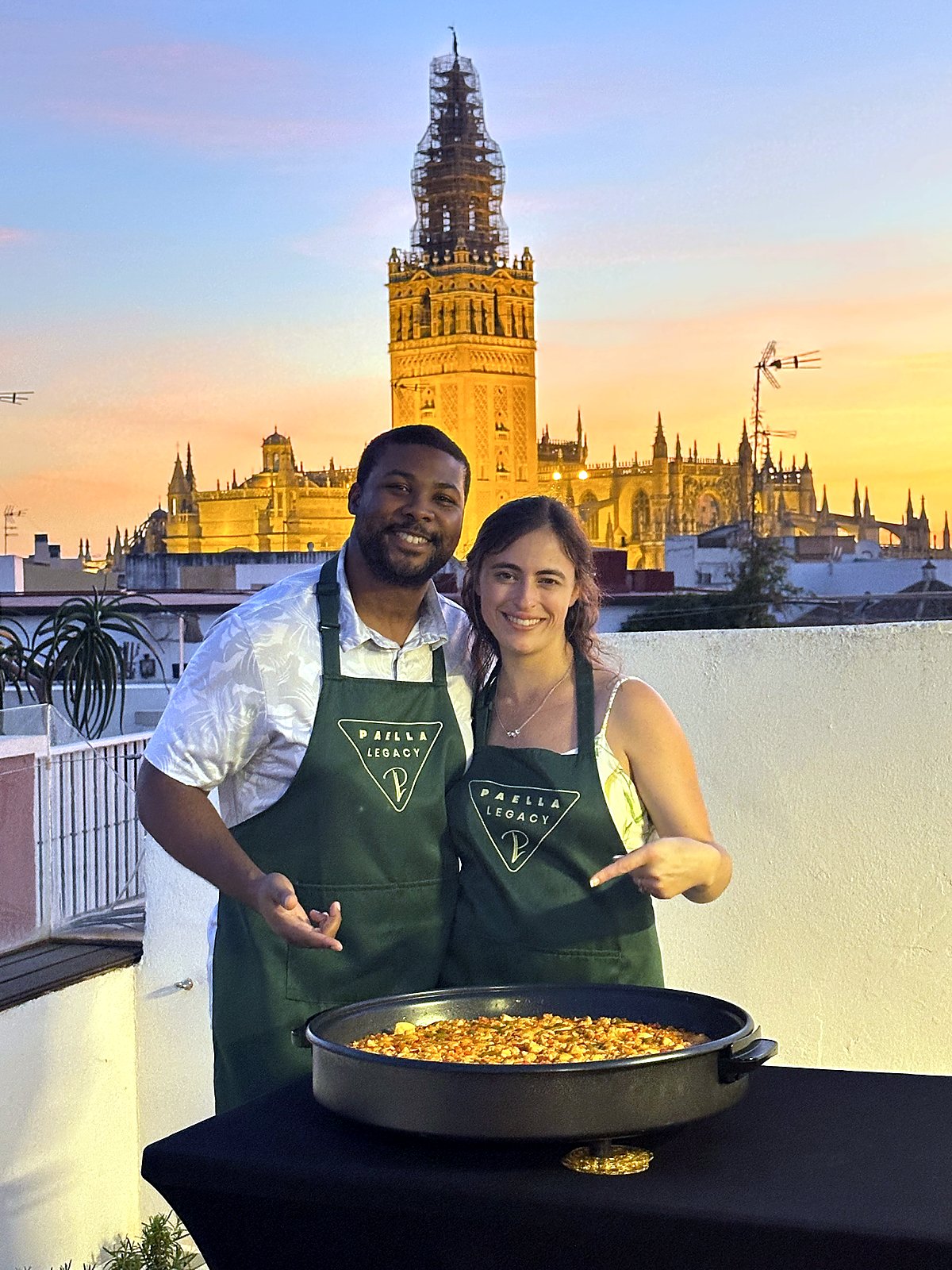 A couple in Paella Legacy aprons with their paella at golden hour, the Giralda and Seville Cathedral lit behind them