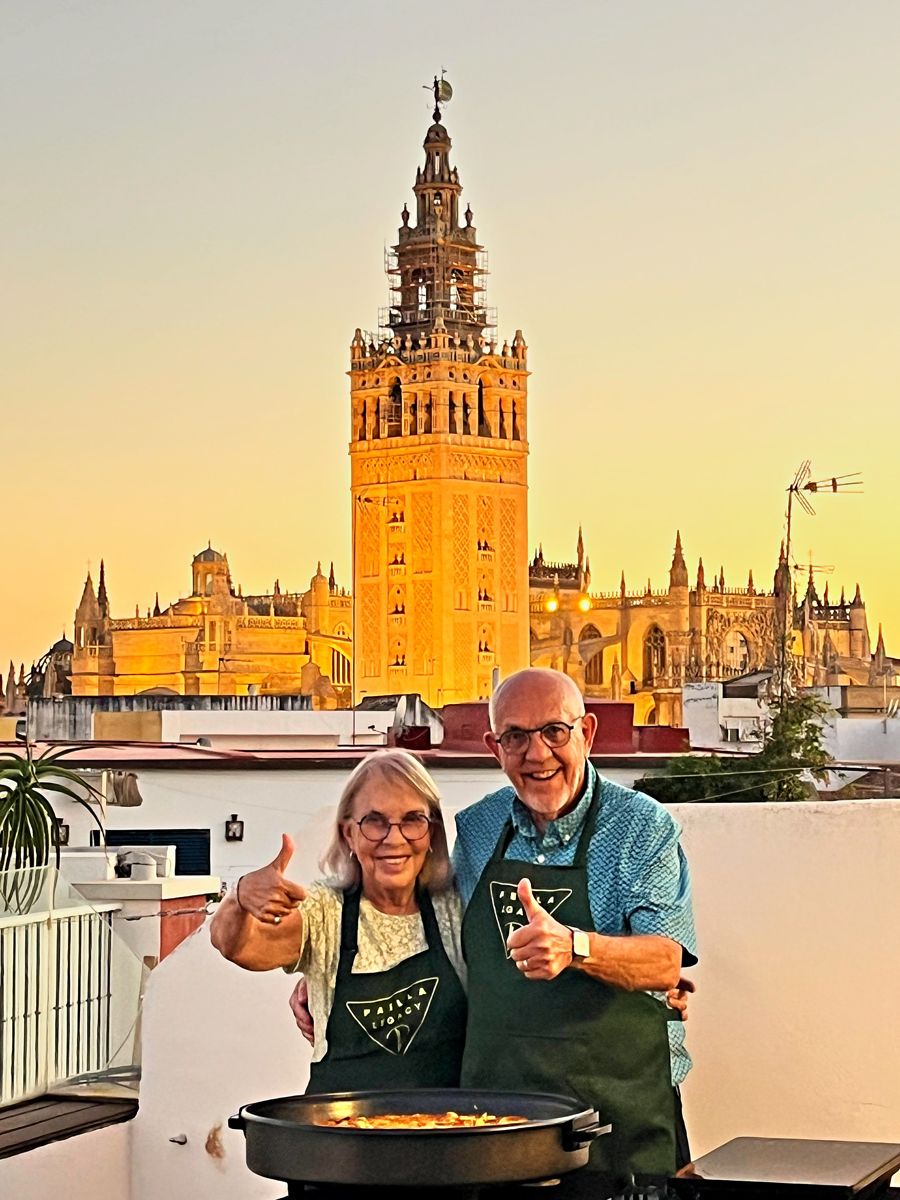 Two guests in Paella Legacy aprons give a thumbs up on the rooftop as the Giralda glows gold at sunset