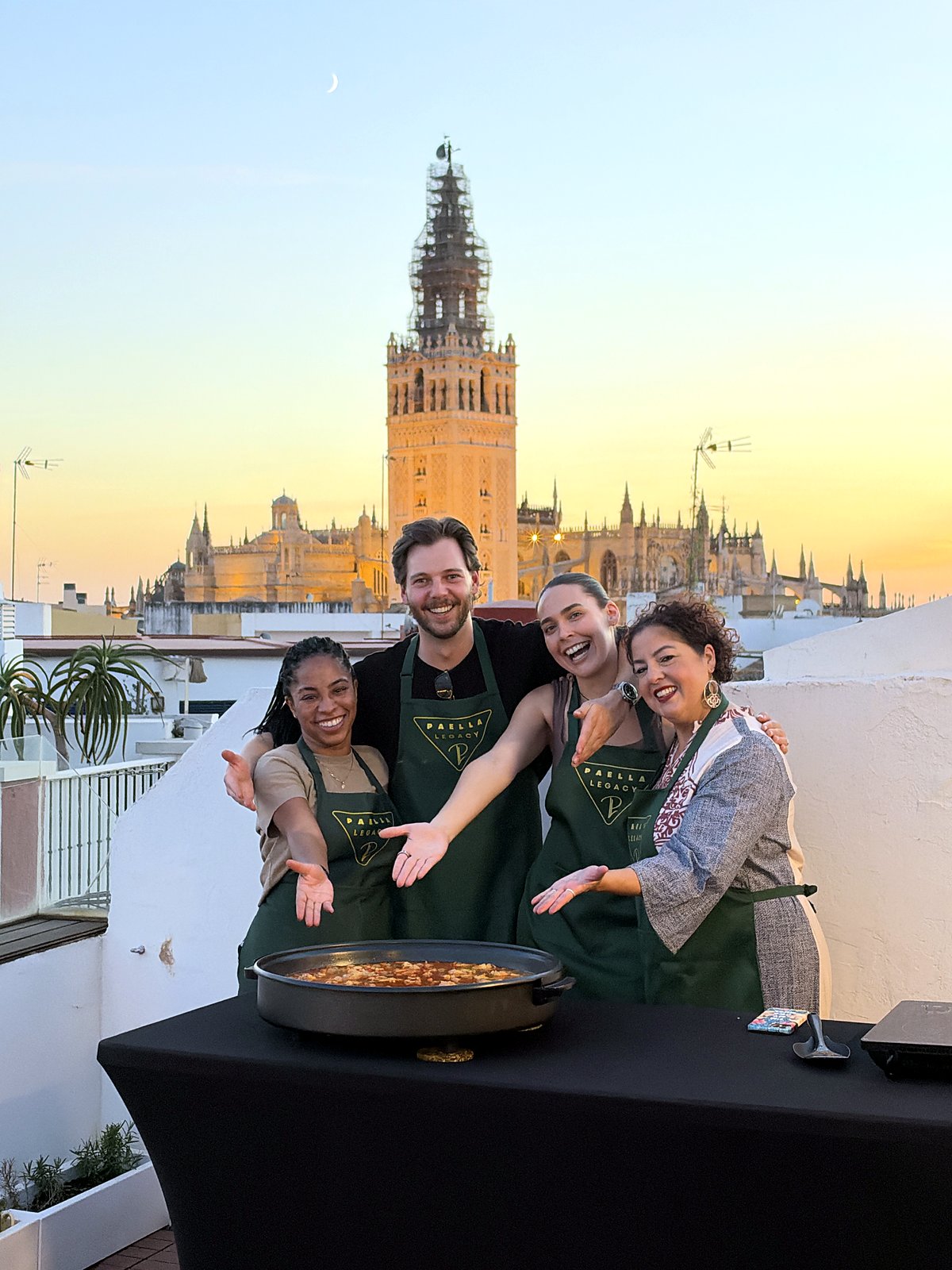Four guests in Paella Legacy aprons laughing beside their finished paella at sunset, the Giralda glowing behind them