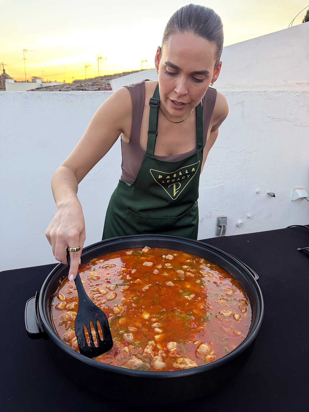A guest in a Paella Legacy apron stirring her paella as the stock simmers, sunset light over the Seville rooftops