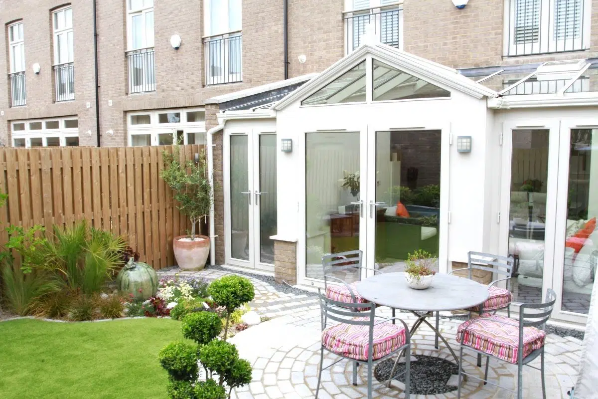Symmetrical white timber-framed conservatory with large French doors opening onto a round mosaic patio, surrounded by landscaped garden featuring decorative dwarf brick walls, greenery, and outdoor seating.