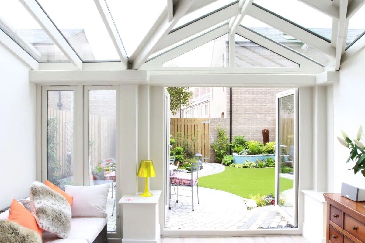 Open plan living-dining conservatory with glass roof, plush seating, and vibrant outdoor garden view featuring green lawn and decorative plants.