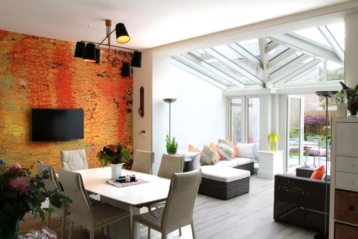 Stylish conservatory interior featuring a gable-end roof, modern dining area with white table and chairs, vibrant textured wall, and abundant natural light from large glass panels.