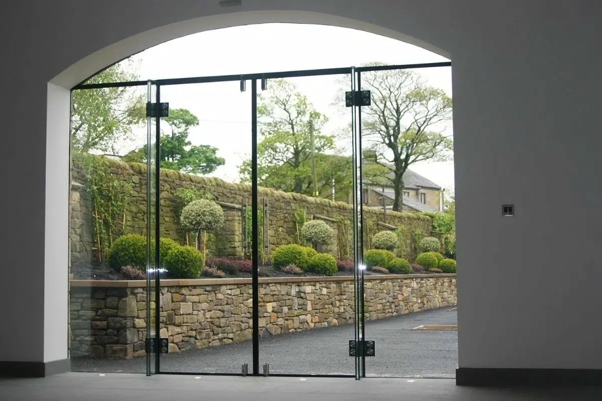 Dramatic arched frameless glass doors opening to a landscaped garden with stone walls and greenery, showcasing seamless integration of modern design and natural surroundings.