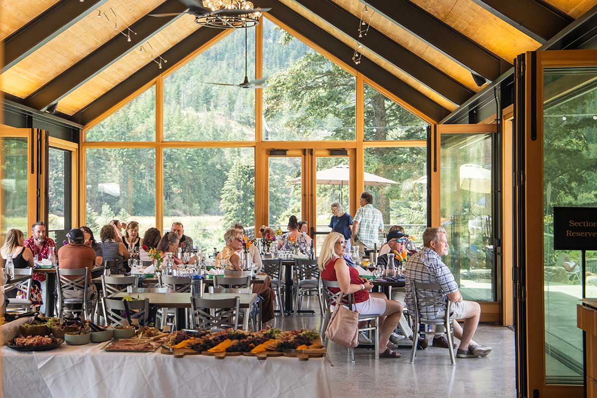 Glass dining pavilion with guests enjoying a meal, surrounded by garden views, featuring large windows, a glass roof, and stylish decor for indoor-outdoor flow.