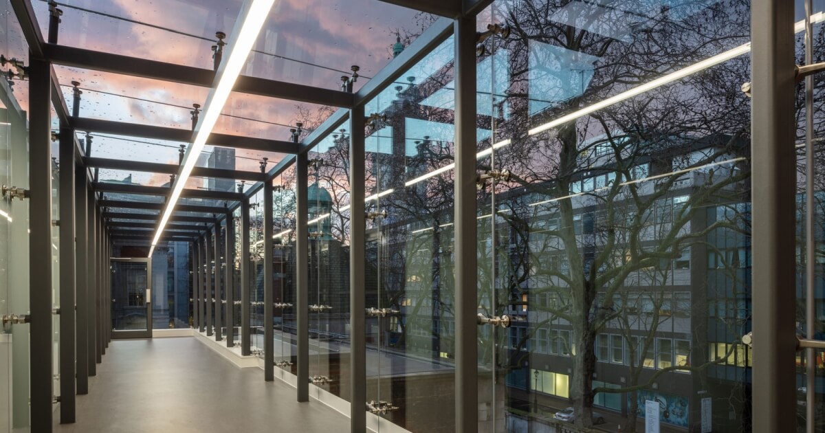 Glass link corridor with transparent walls, framed by structural supports, showcasing modern design and natural light, enhancing connectivity between buildings.