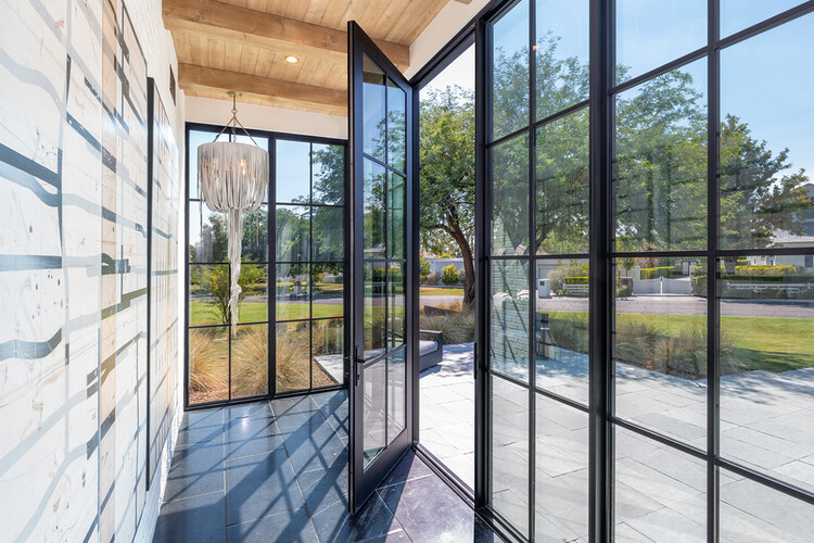 Glass entrance hall with open doors, featuring structural glass walls, a modern chandelier, and views of an outdoor garden, emphasizing transparency and connection to nature.