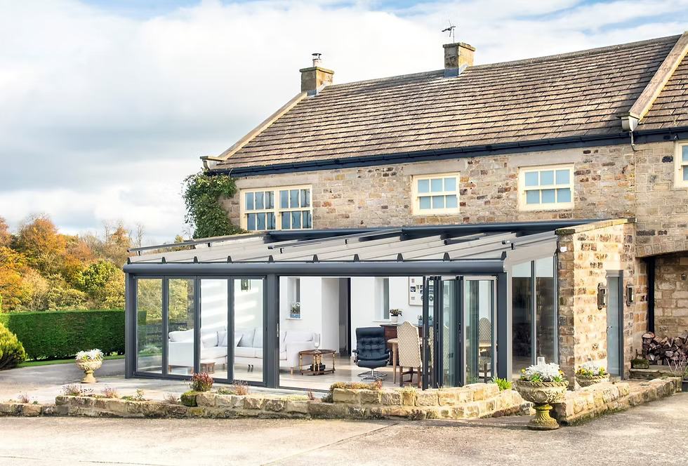 Aluminium conservatory with floor-to-ceiling glazing, ultra-slim profiles, and a modern design, enhancing natural light and garden views, adjacent to a stone house.