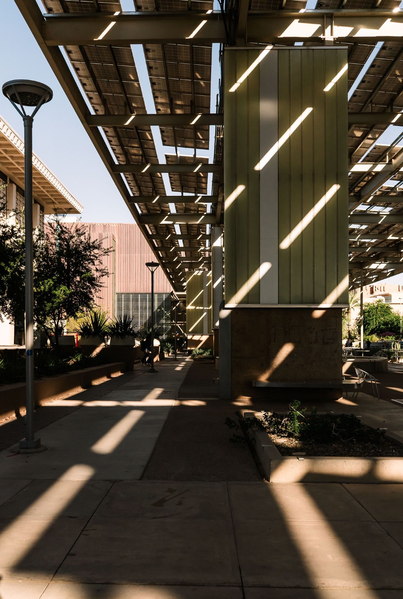 Modern outdoor walkway with solar panel canopy casting geometric shadows across a landscaped plaza.