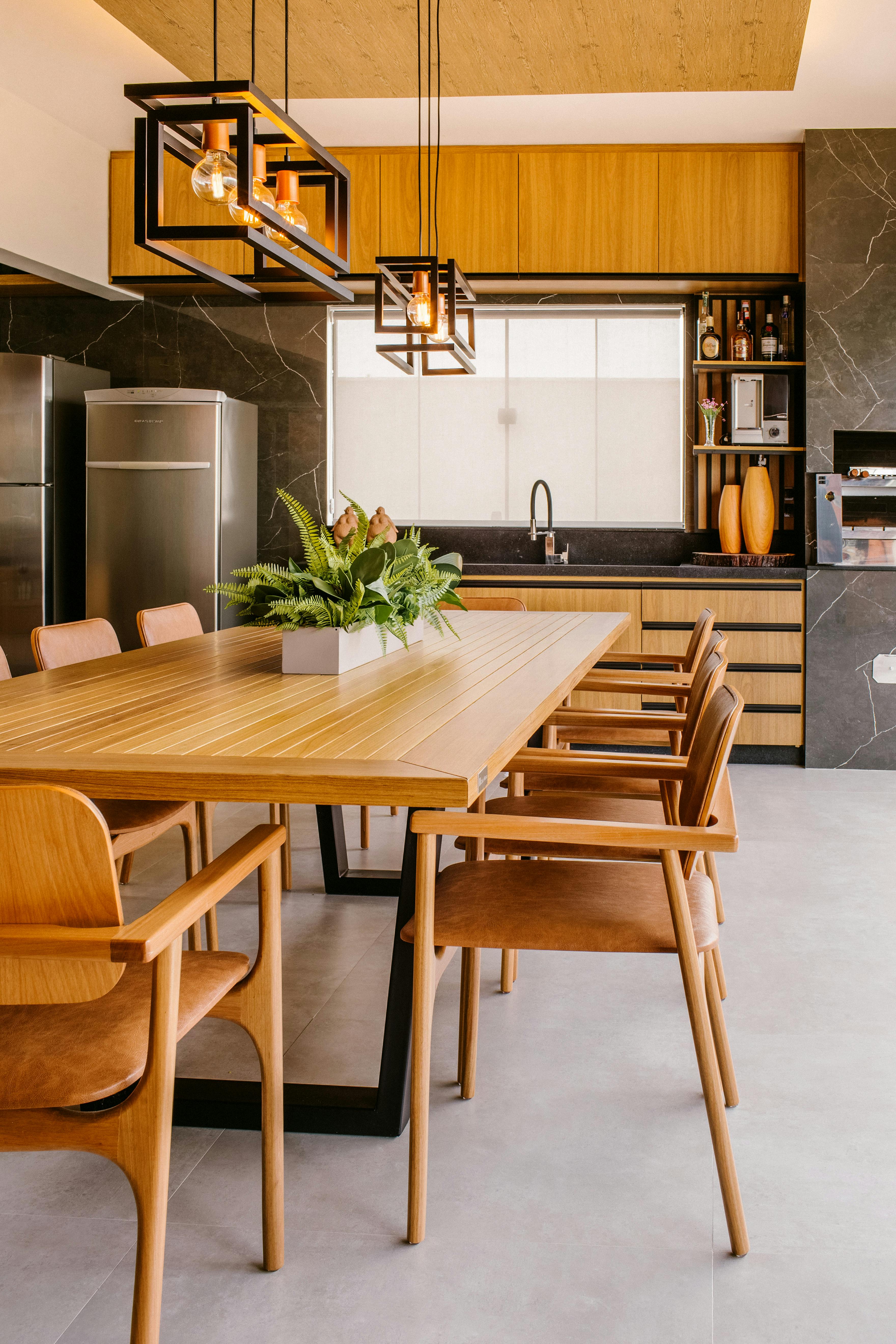 Modern luxury kitchen and dining area with wood cabinetry, black marble backsplash, minimalist pendant lighting, and a large wooden dining table with upholstered chairs in a contemporary open-concept home.