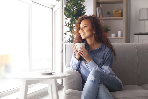 Woman with curly hair sitting on a sofa by a bright window, holding a mug and looking outside.