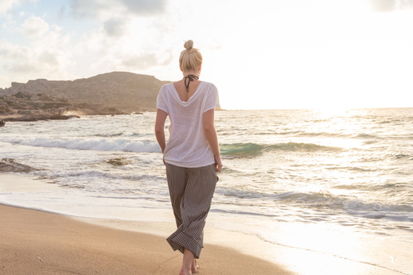 Woman walking barefoot along a beach toward the light, seen from behind.