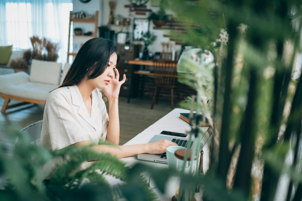 A woman with long dark hair sitting at a desk with her laptop, resting her head against her hand and looking down in thought, with green plants in the foreground. A woman with long dark hair sitting at a desk with her laptop, resting her head against her hand and looking down in thought, with green plants in the foreground.