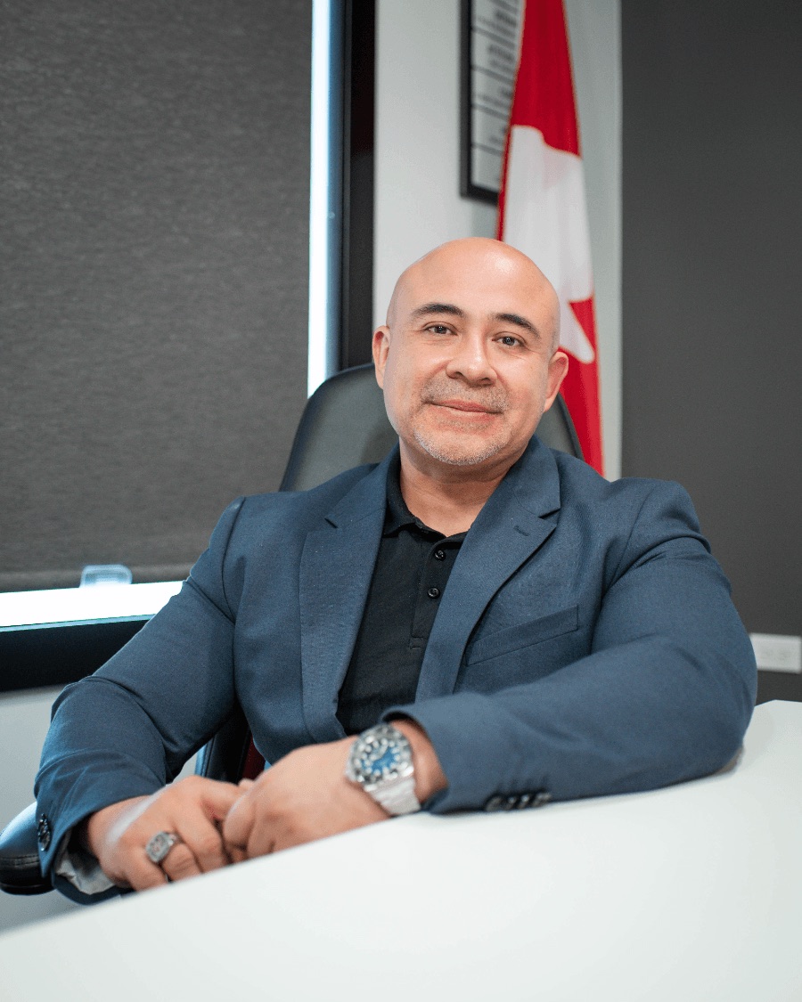 Hector Alvarado seated at his desk in a navy blazer, with a Canadian flag behind him