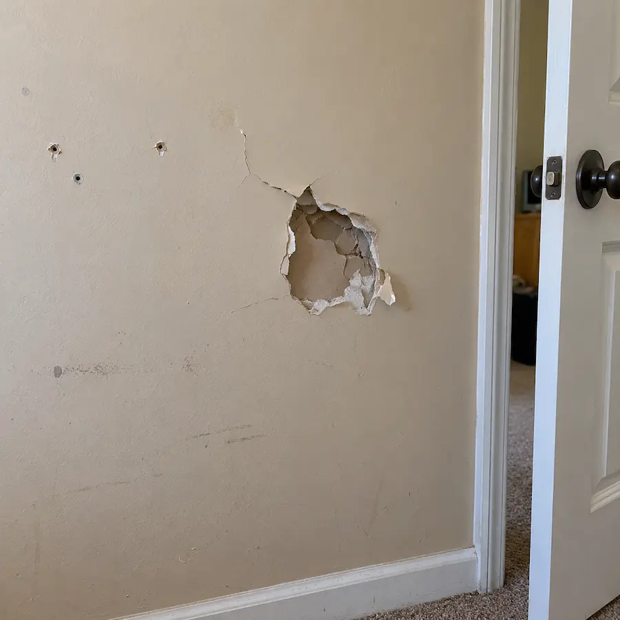 Doorknob-sized hole with torn paper and cracks in a beige drywall wall, with anchor holes beside it