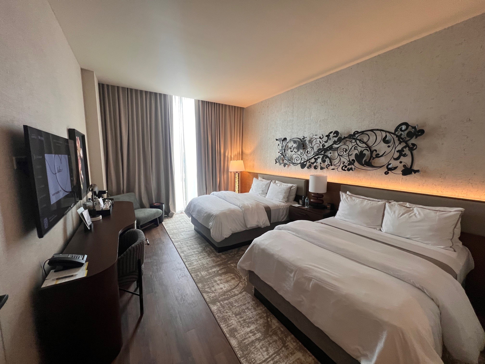 Renovated double queen guest room at the Westin Nashville with two white queen beds, decorative black scrollwork wall art with warm amber uplighting, hardwood floors, floor-to-ceiling curtained windows, a flat-screen TV, and a work desk with chair