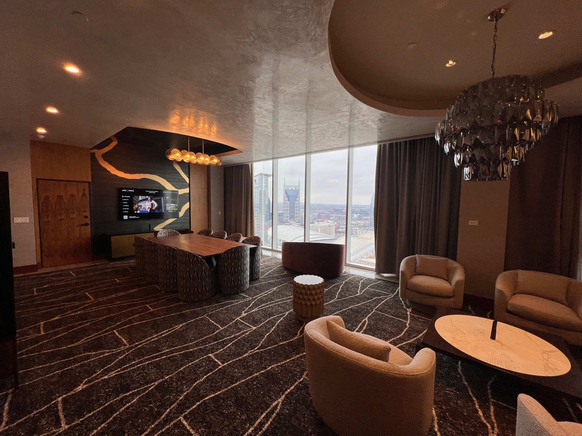 Renovated Broadway Suite living room at the Westin Nashville with panoramic floor-to-ceiling windows showcasing the Nashville skyline including the AT&T Batman Building, a large wood conference table, sculptural lounge chairs, a bold gold-veined black accent wall with flat-screen TV, dark patterned carpet, and a dramatic smoked-crystal chandelier