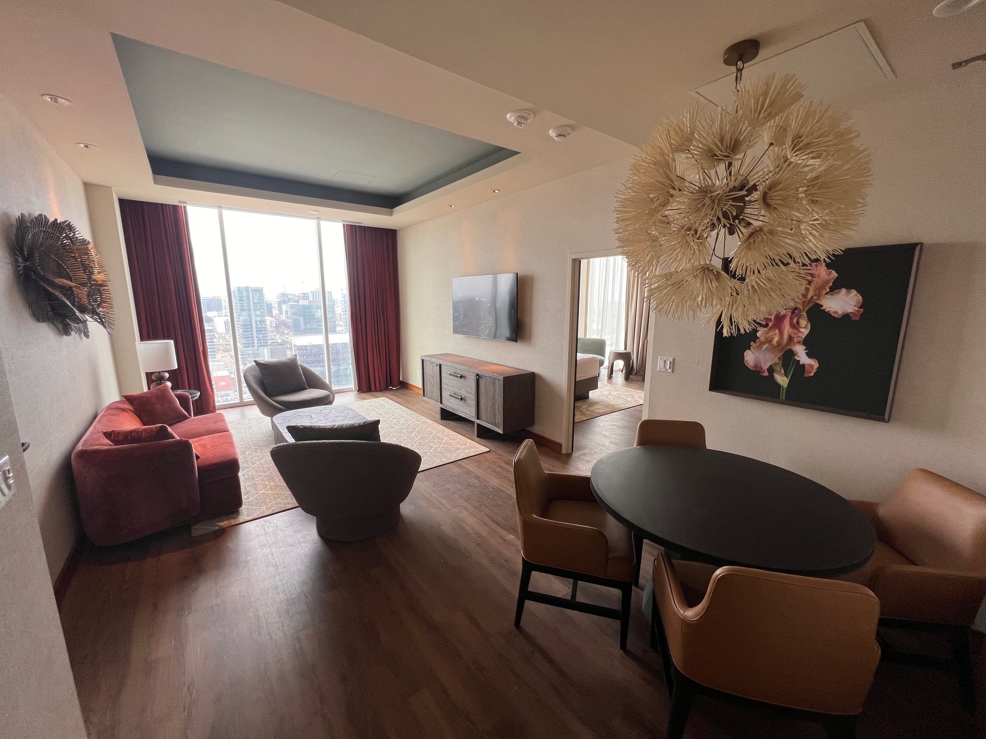 Renovated Executive Suite living and dining area at the Westin Nashville with a round black dining table and tan leather chairs, burgundy velvet sofa, sculptural pendant chandelier, floor-to-ceiling windows overlooking the Nashville skyline, wall-mounted flat-screen TV, and bold floral canvas artwork