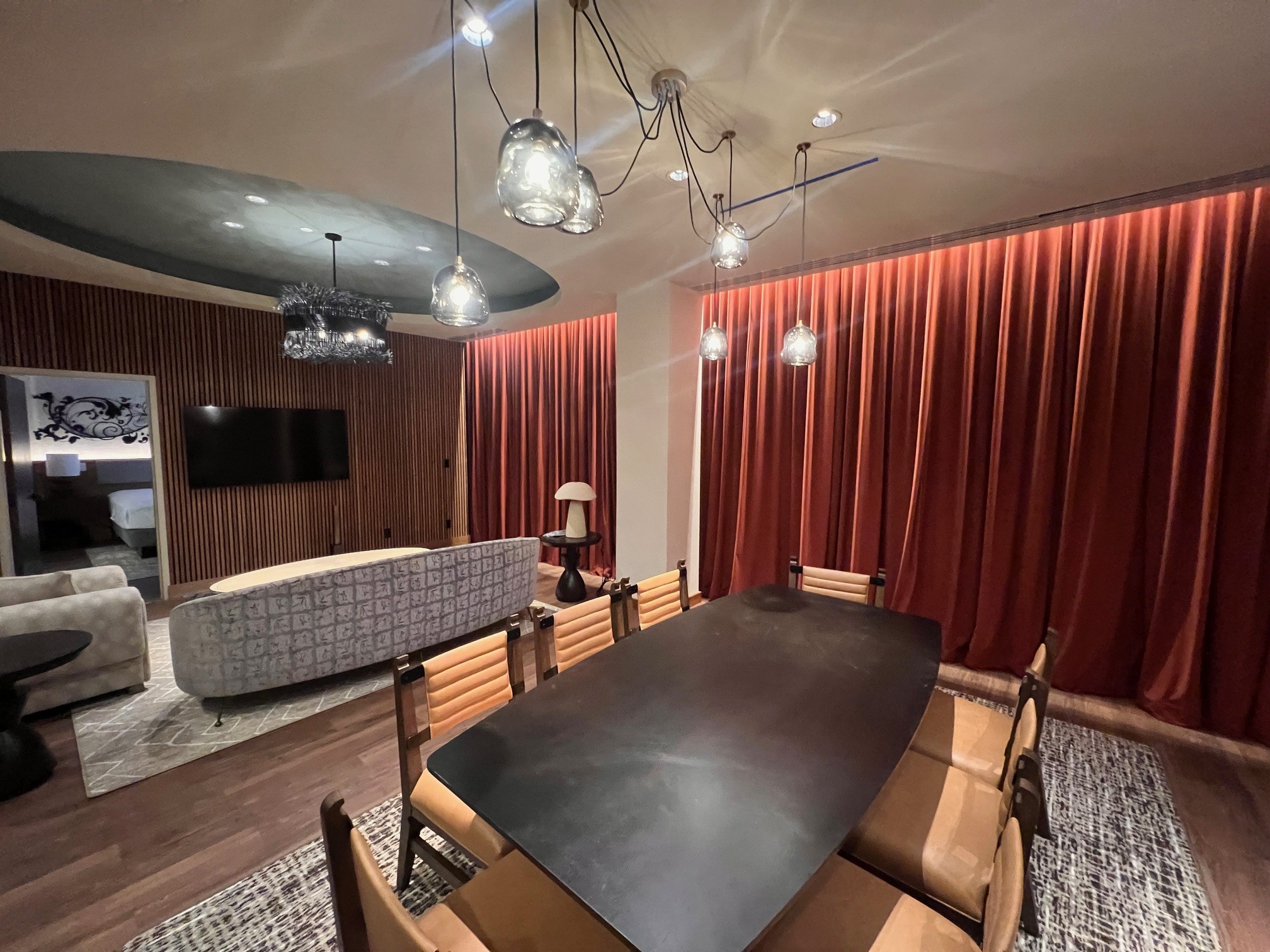 Renovated Belmont Suite dining and living area at the Westin Nashville featuring a large black rectangular dining table with tan leather chairs, sculptural glass pendant lights, a curved patterned sofa, slatted wood accent wall with flat-screen TV, and dramatic floor-to-ceiling rust-red velvet drapes