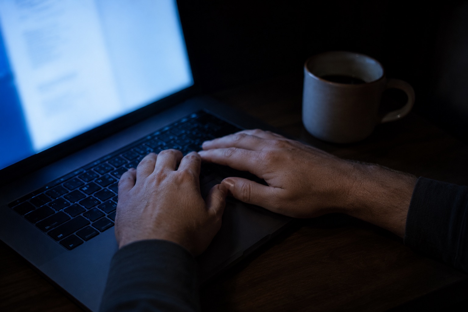 A man's hands on a laptop keyboard late at night, lit only by the screen.