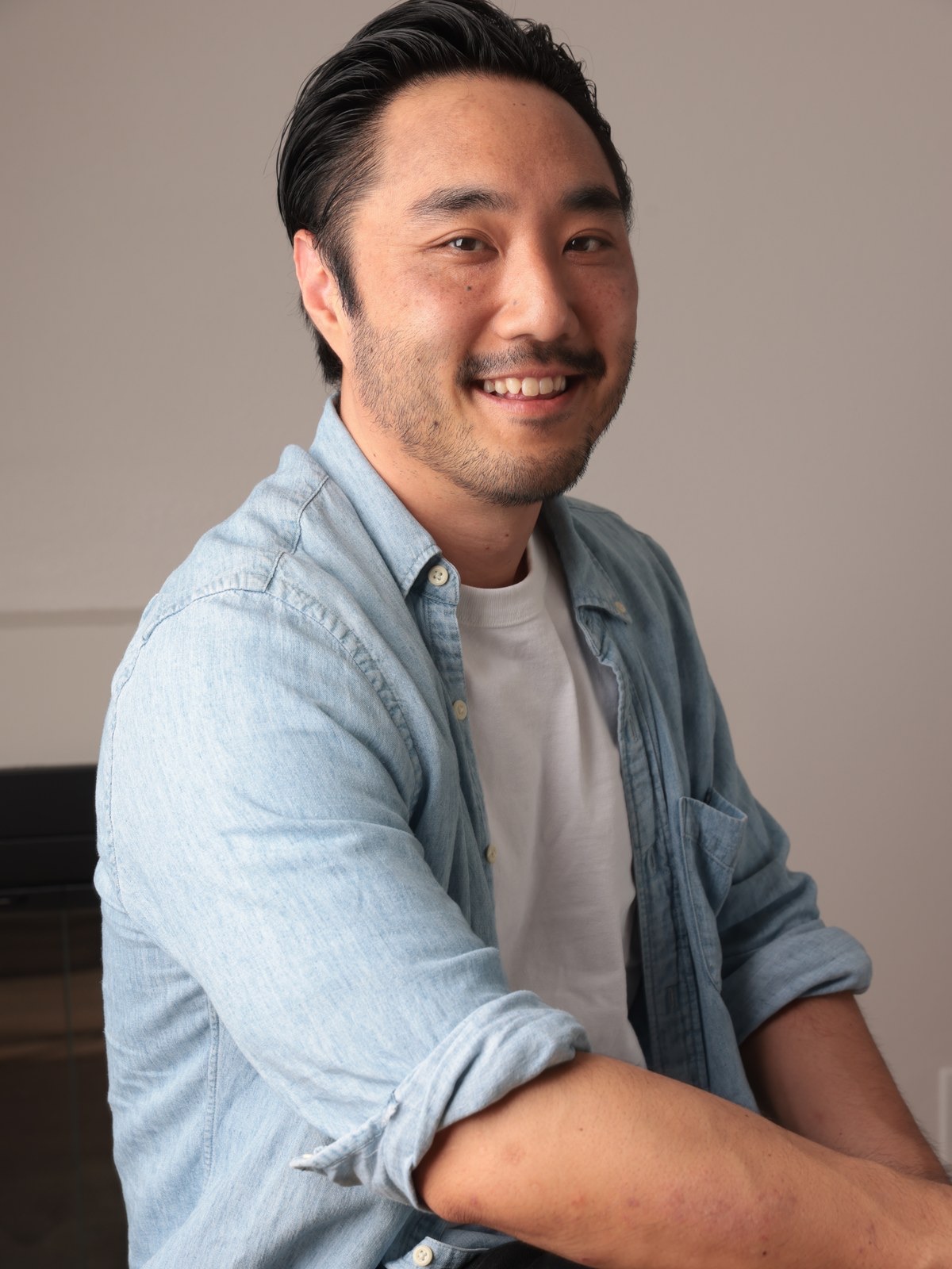 Jeff Kimura seated on a stool in a bright room, chambray shirt over a white tee, looking off-camera mid-conversation.