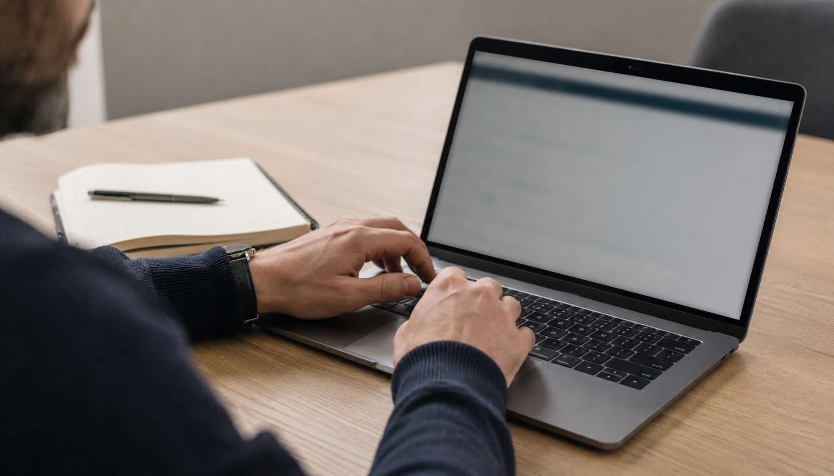 A person typing on a laptop at a wooden desk, an open notebook and pen beside it.