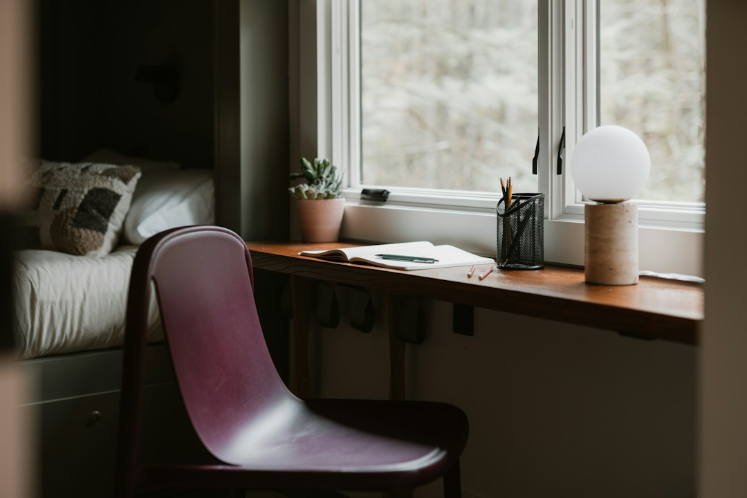 A wooden desk shelf beneath a bright window with an open notebook, pencil cup, small plant and a round lamp beside a chair.