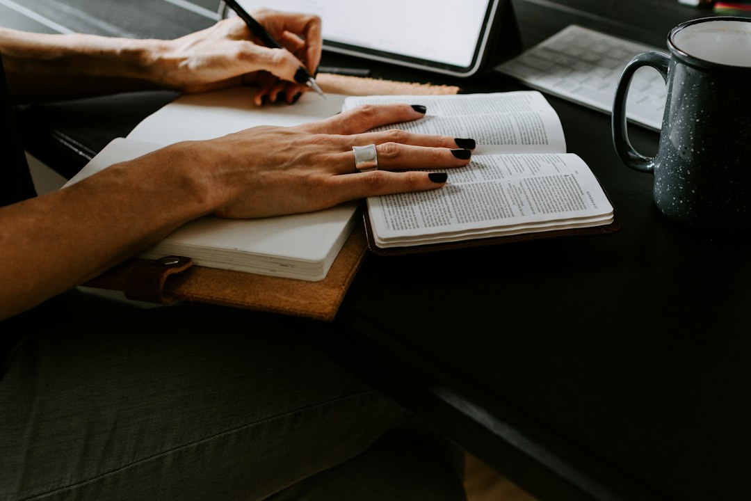 Close view of a person's hands resting on an open book and notebook on a dark table beside a speckled enamel mug.