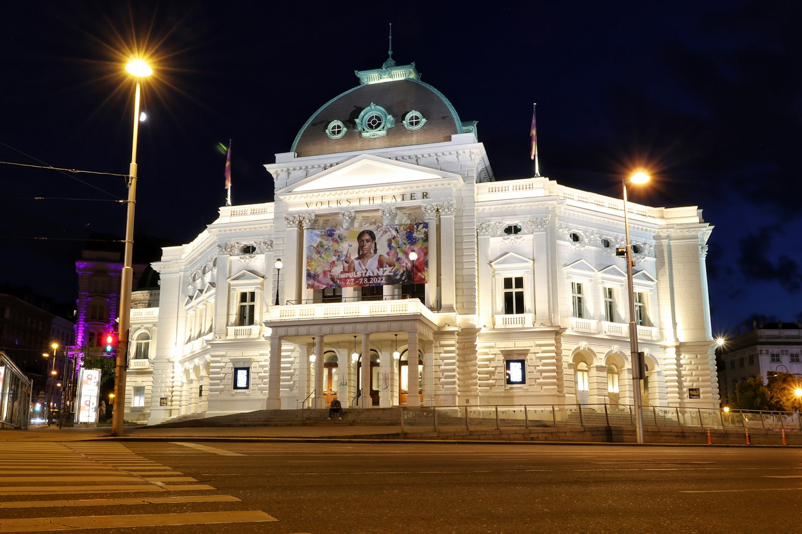 The illuminated white Volkstheater in Vienna at night, with a green dome and street lamps in the foreground