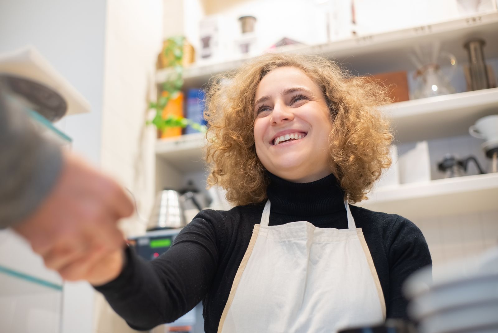 A smiling cafe owner in an apron shaking hands with a returning customer across the counter