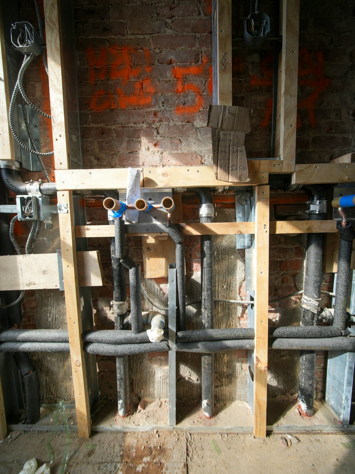 Insulated plumbing pipes and electrical conduit exposed inside a brick wall, illustrating the building systems behind maintenance requests