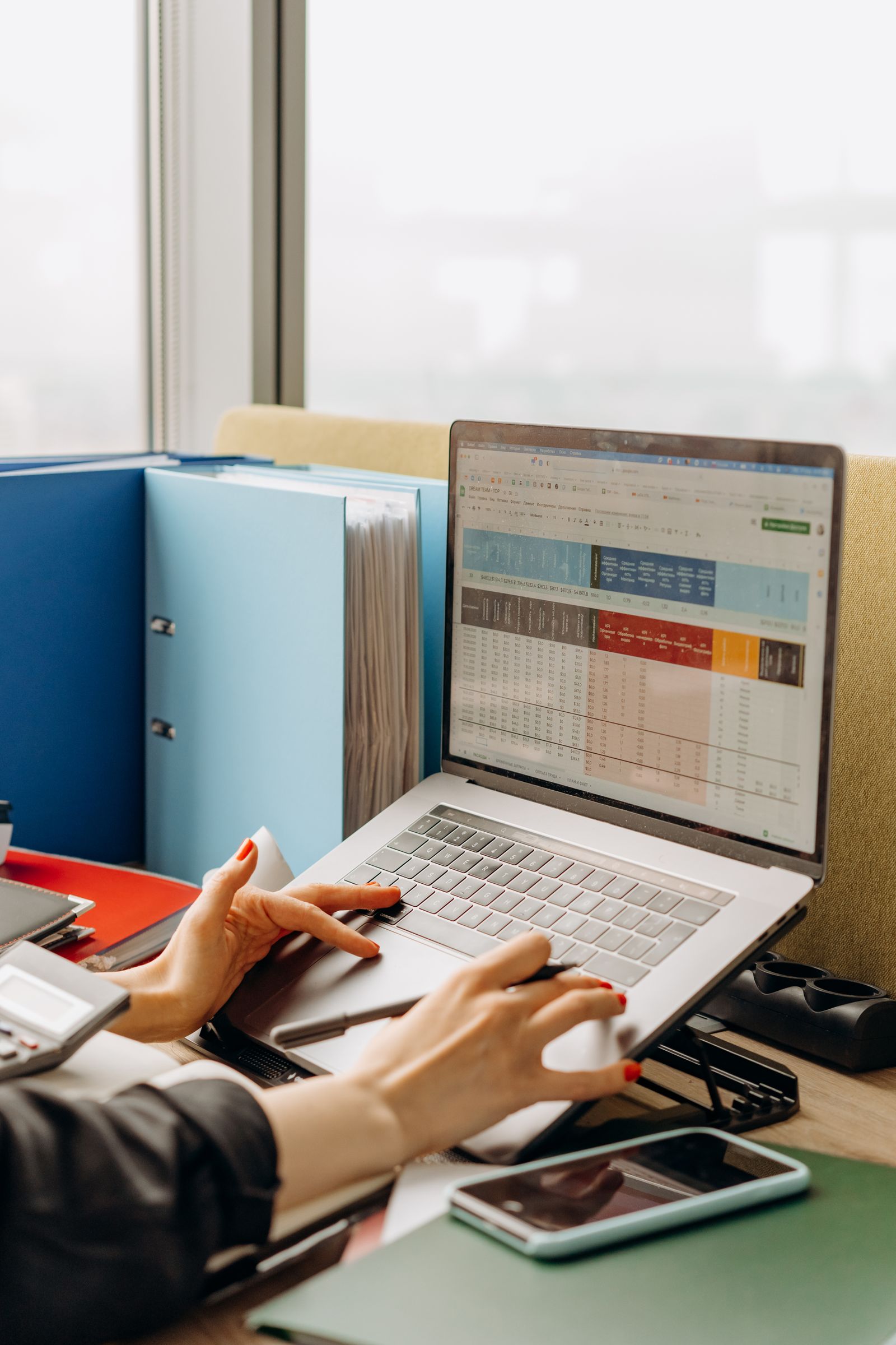 A person working through a customer spreadsheet on a laptop beside binders and a smartphone