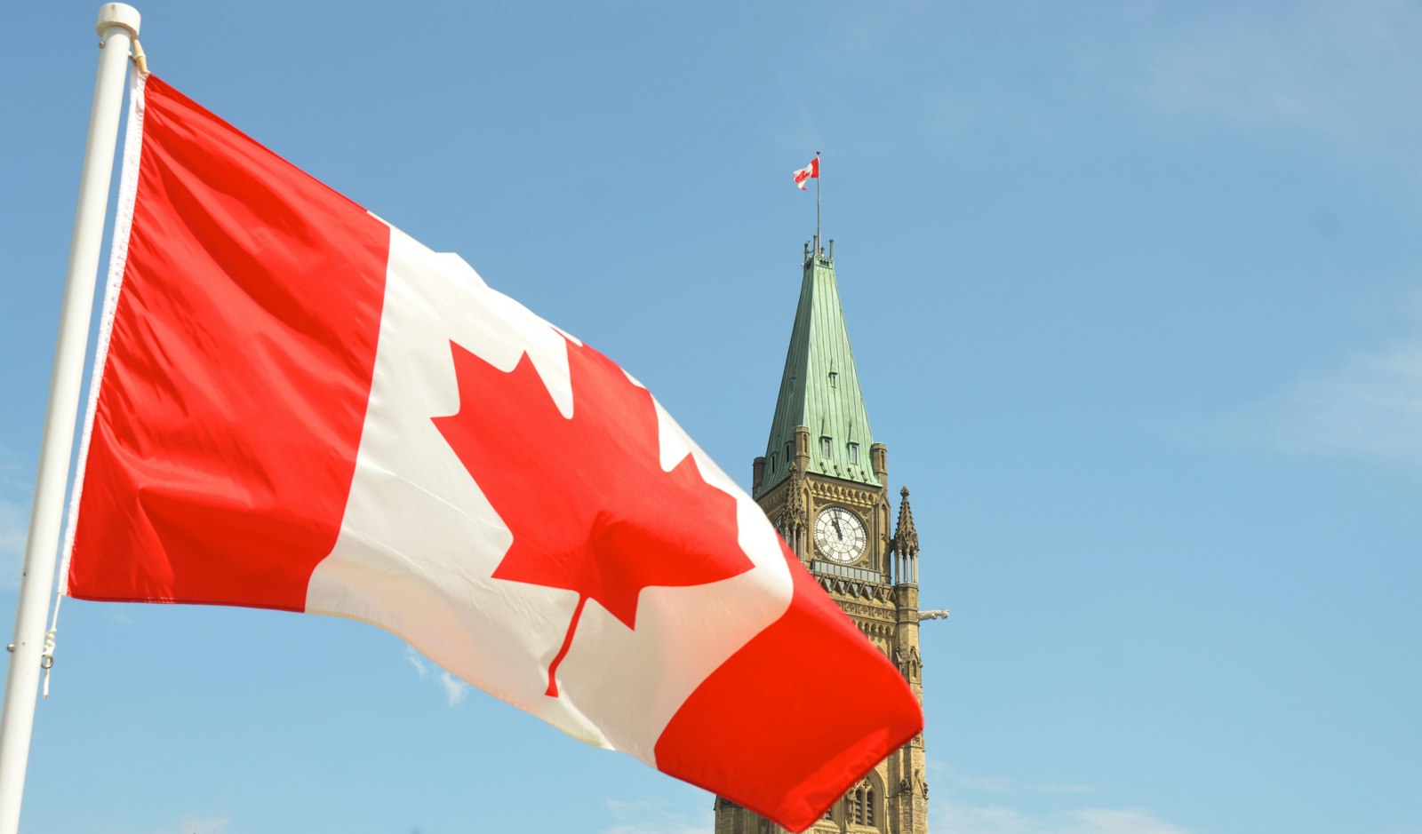 A Canadian flag waving in front of the Peace Tower on Parliament Hill in Ottawa under a clear blue sky
