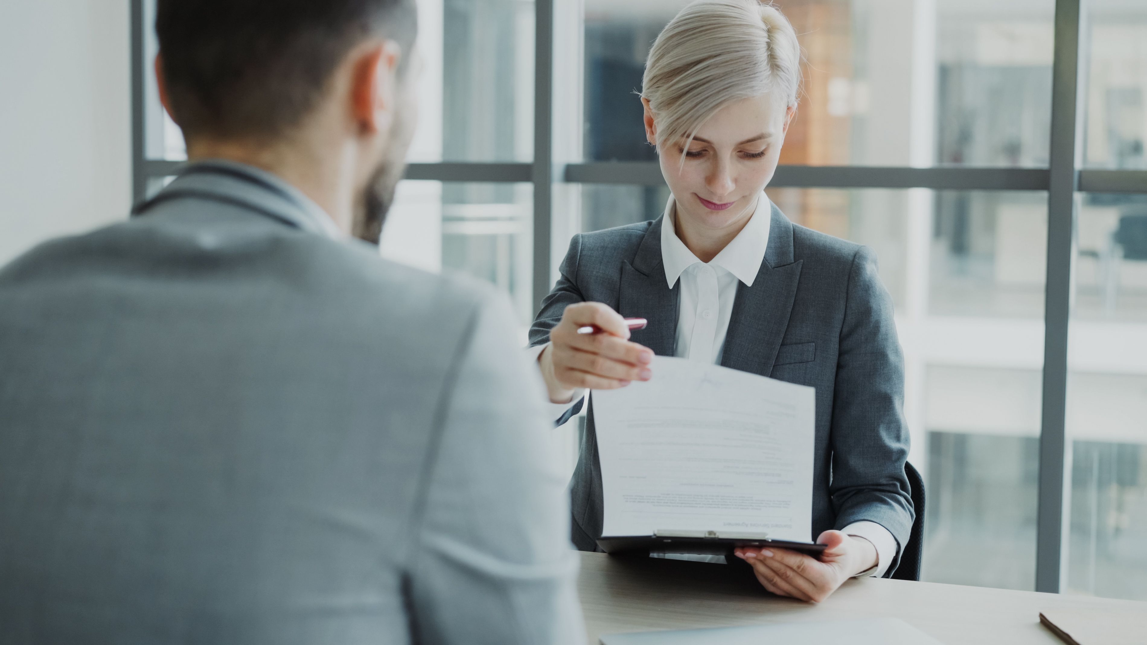 Two colleagues at an office table review and hand over a document on a clipboard