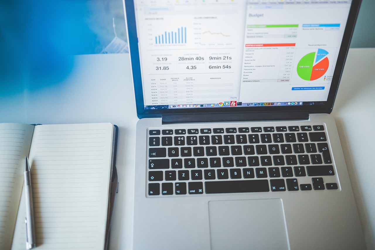 Close-up of a laptop screen displaying a budget dashboard with a bar chart, pie chart, and expense figures next to an open notebook